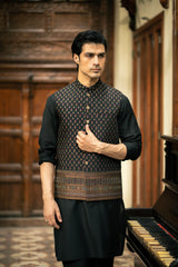 Man wearing a traditional black kurta with a patterned vest in a room with wooden paneling and a piano.