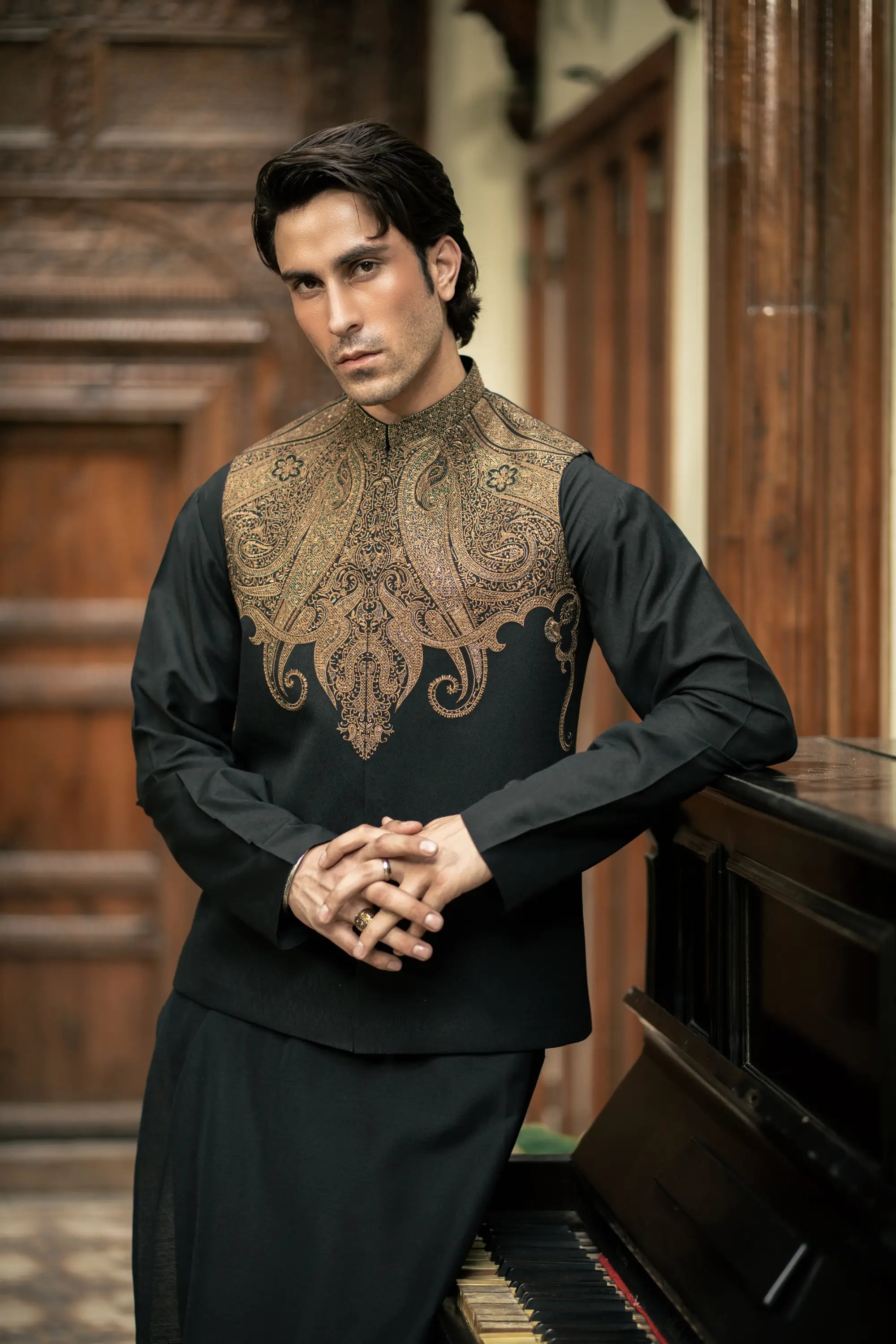 Man wearing a black traditional outfit with intricate gold embroidery in an indoor setting.