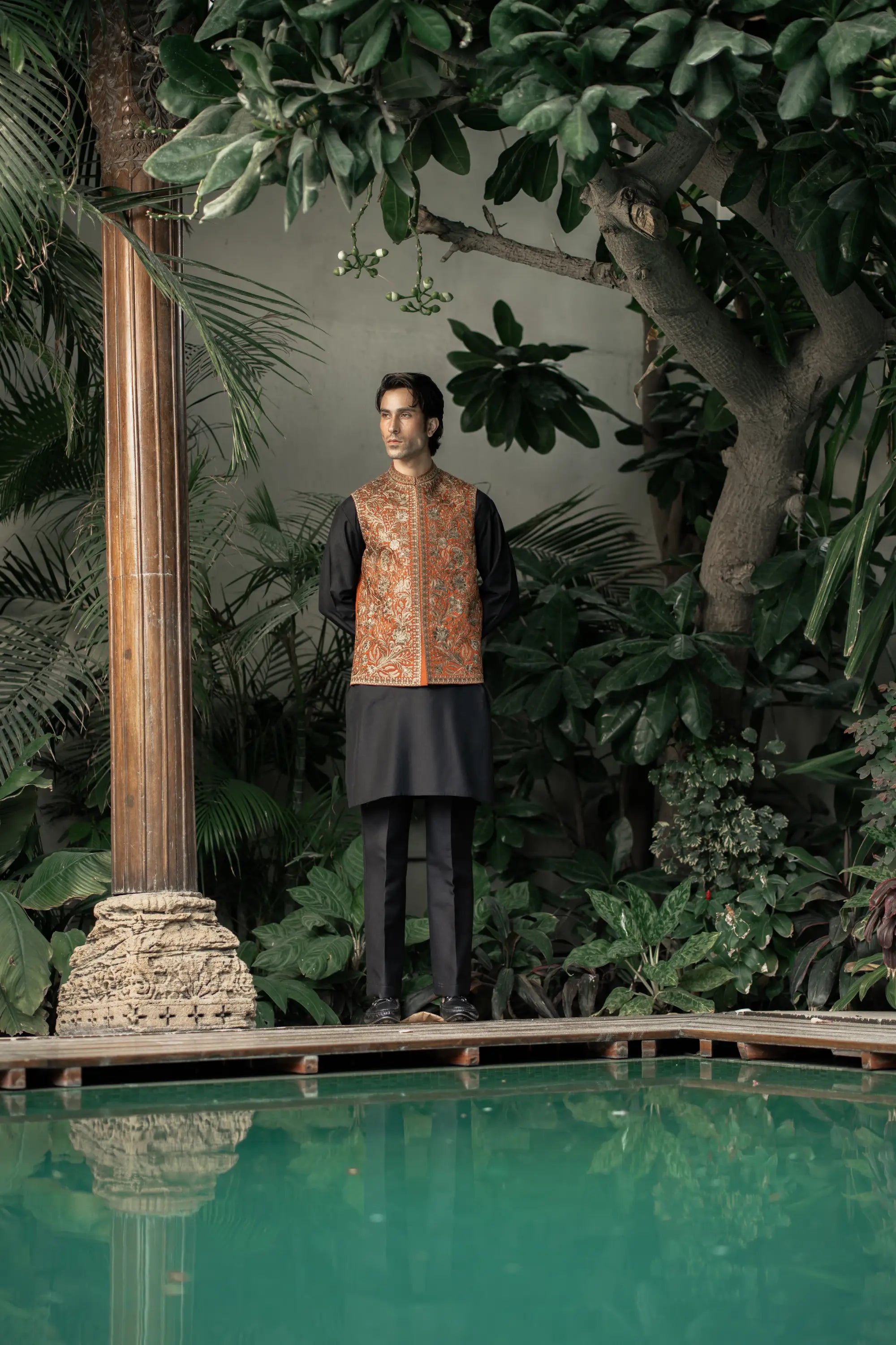 Man in traditional outfit standing by a pool with lush greenery
