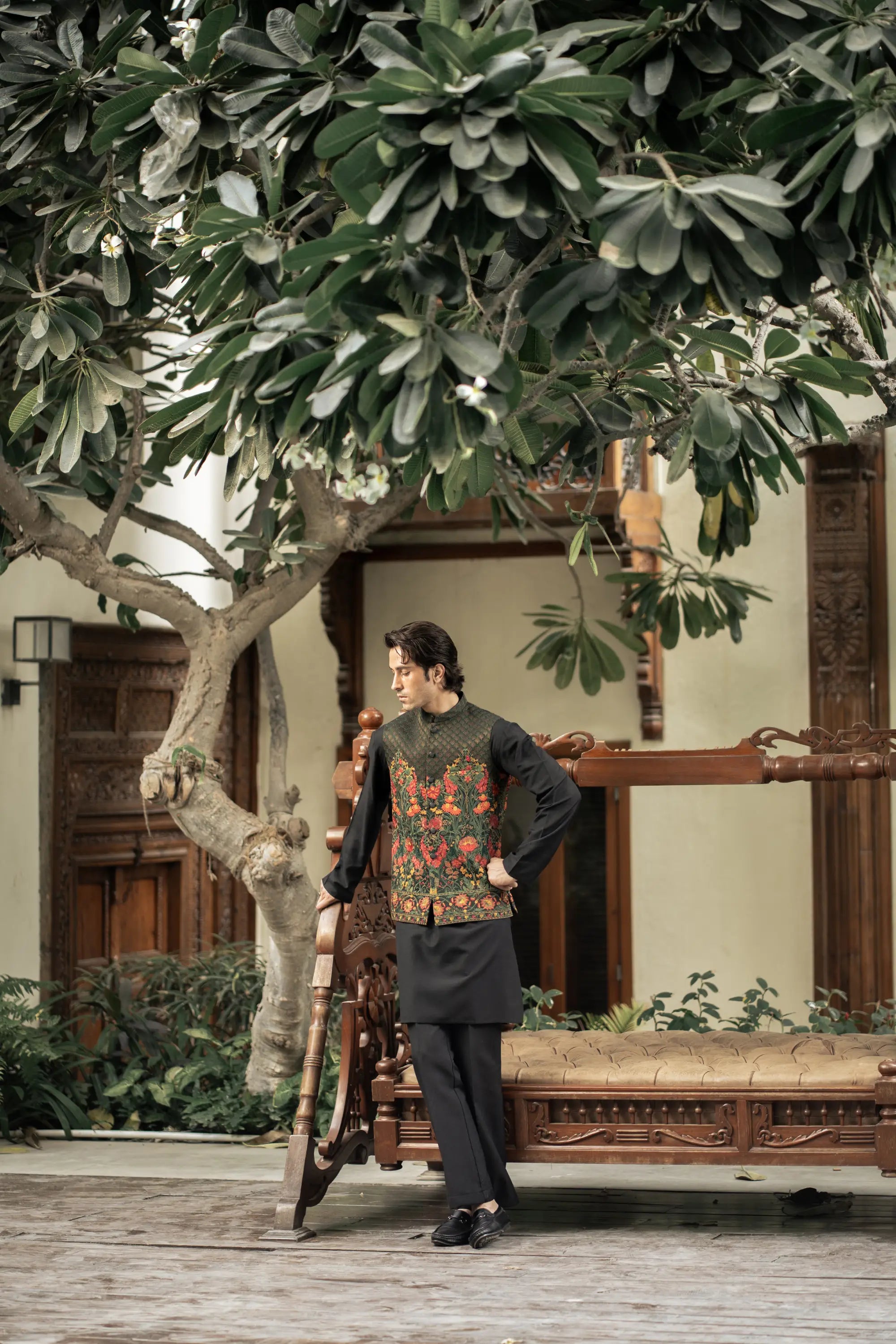 Man standing under a large tree in an outdoor setting with wooden furniture and plants.