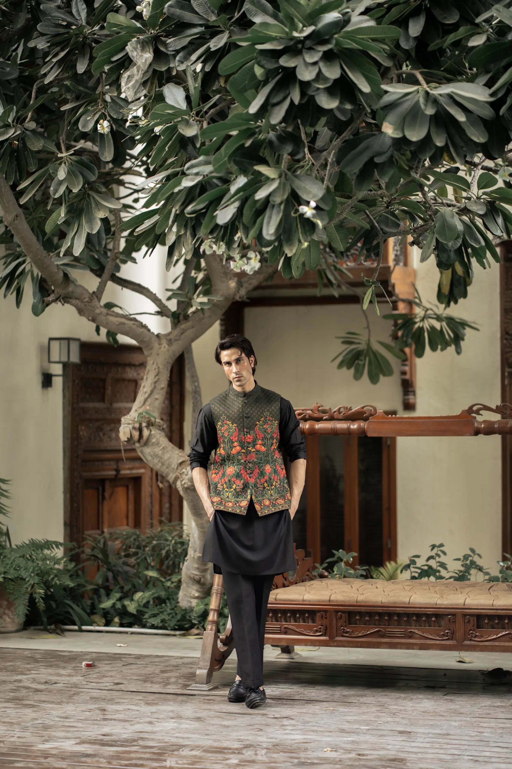 Man standing in a garden setting with a tree and traditional furniture.