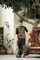Man wearing a traditional outfit standing in an outdoor setting with plants and a wooden chair.
