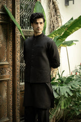 Man wearing a black traditional outfit standing in front of an ornate wooden door with plants in the background.