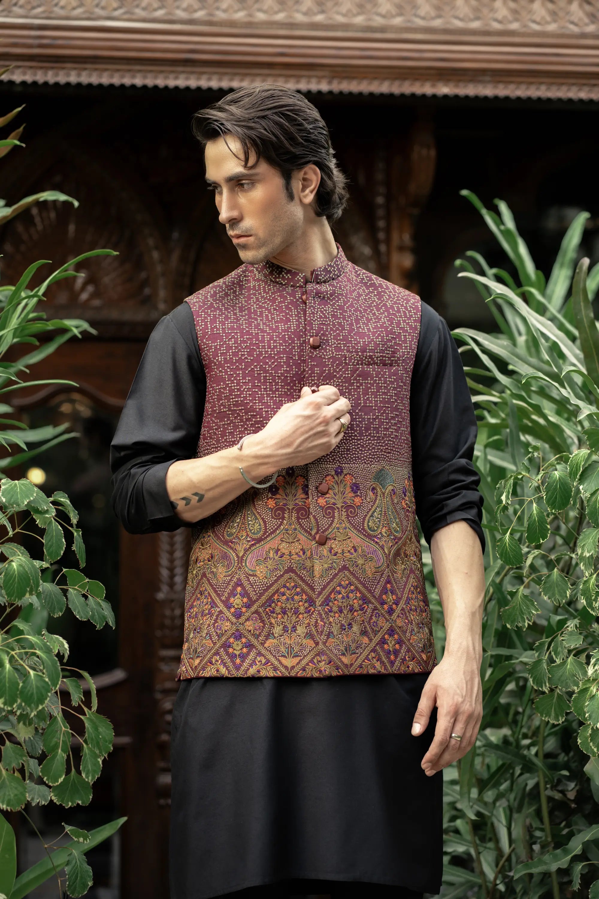 Man wearing a traditional outfit with a patterned vest and black kurta in a decorative setting.