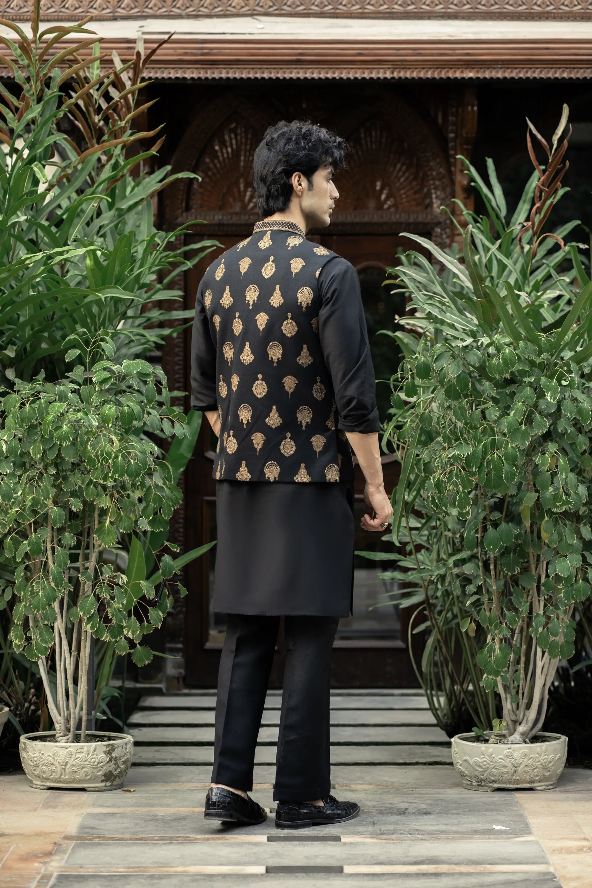 Man wearing a black kurta with a patterned jacket standing in front of a building with plants.
