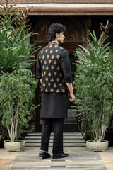 Man wearing a black kurta with a patterned jacket standing in front of a building with plants.