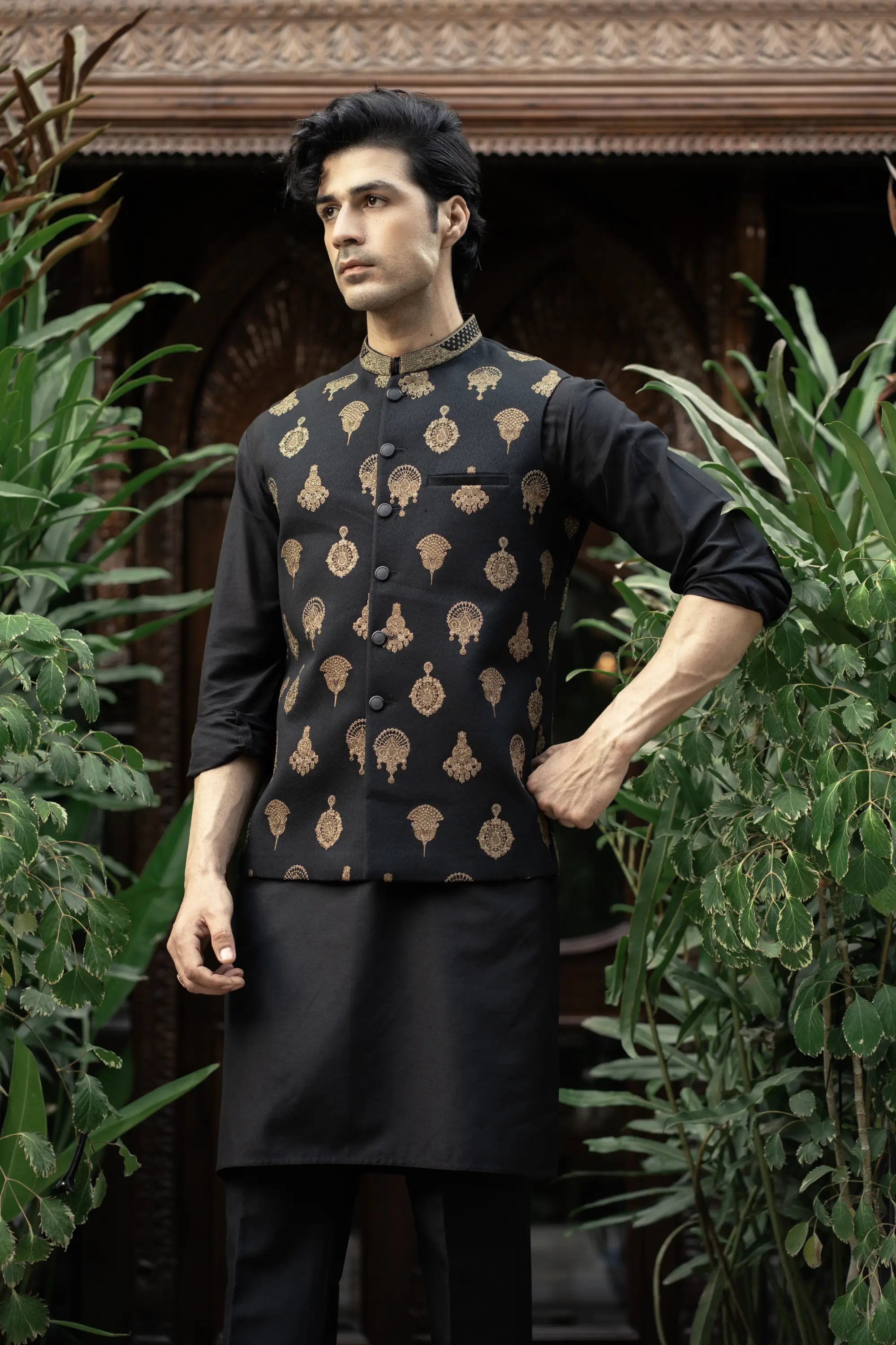 Man wearing a black kurta with gold patterns standing among green plants.