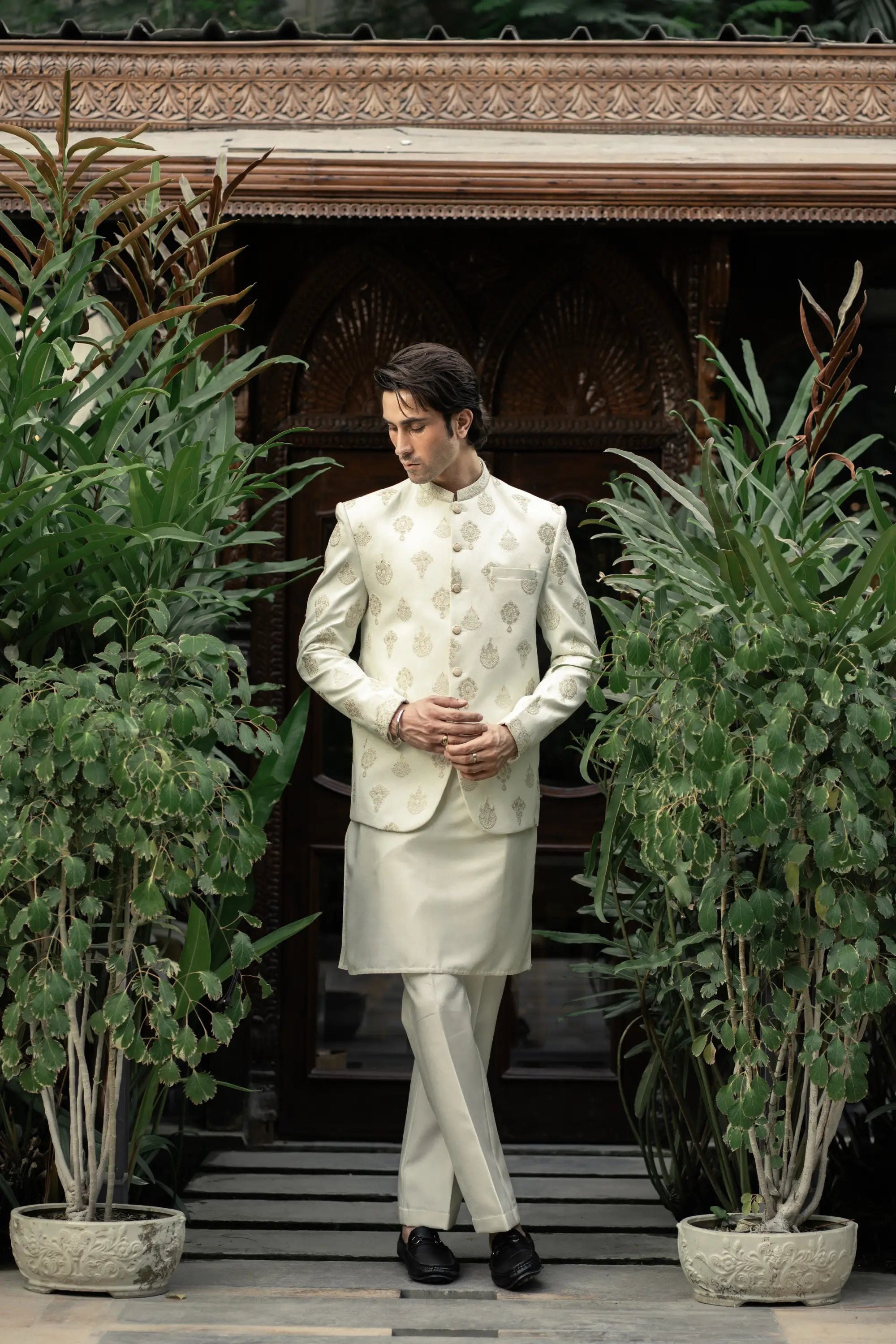 Man in a traditional white sherwani standing in front of decorative wooden panels with plants around.