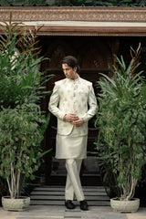 Man in a traditional white sherwani standing in front of decorative wooden panels with plants around.