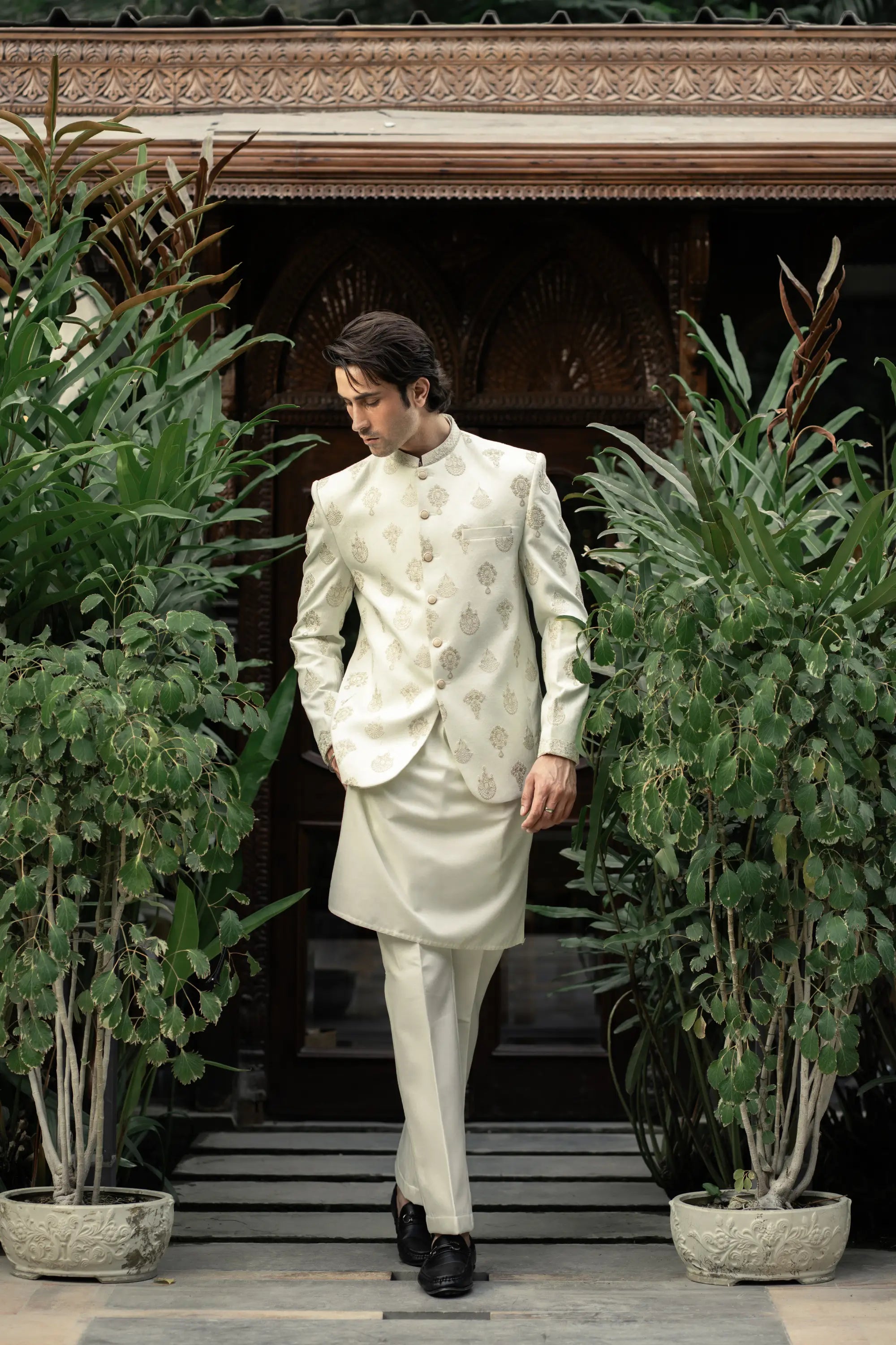 Man in a traditional white sherwani standing between potted plants.