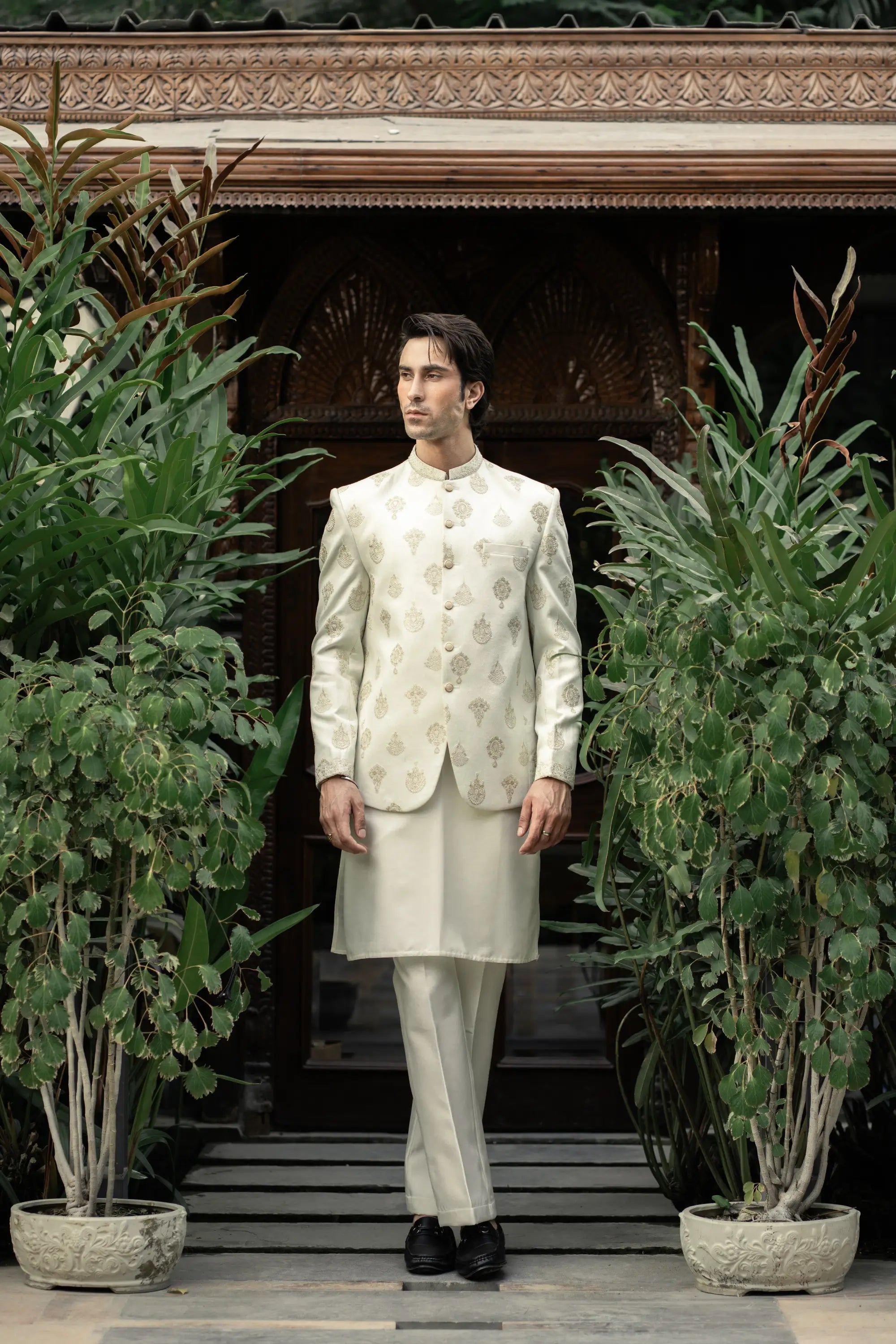 Man in a traditional white outfit standing in front of a decorative entrance with plants.