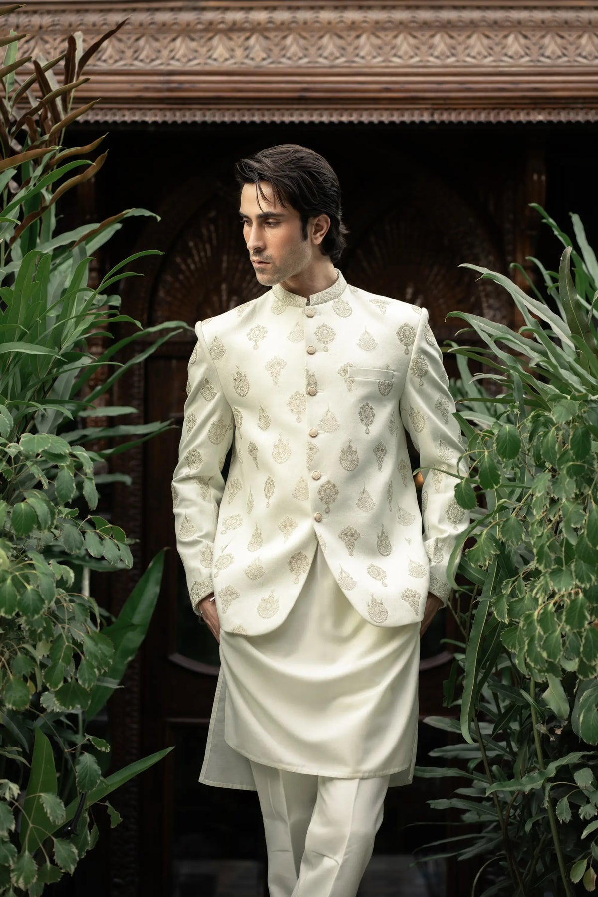 Man wearing a traditional white sherwani with intricate patterns in a garden setting