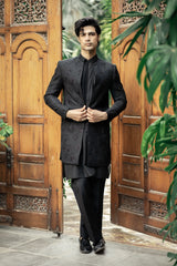 Man in a black traditional outfit standing in front of wooden doors with greenery.