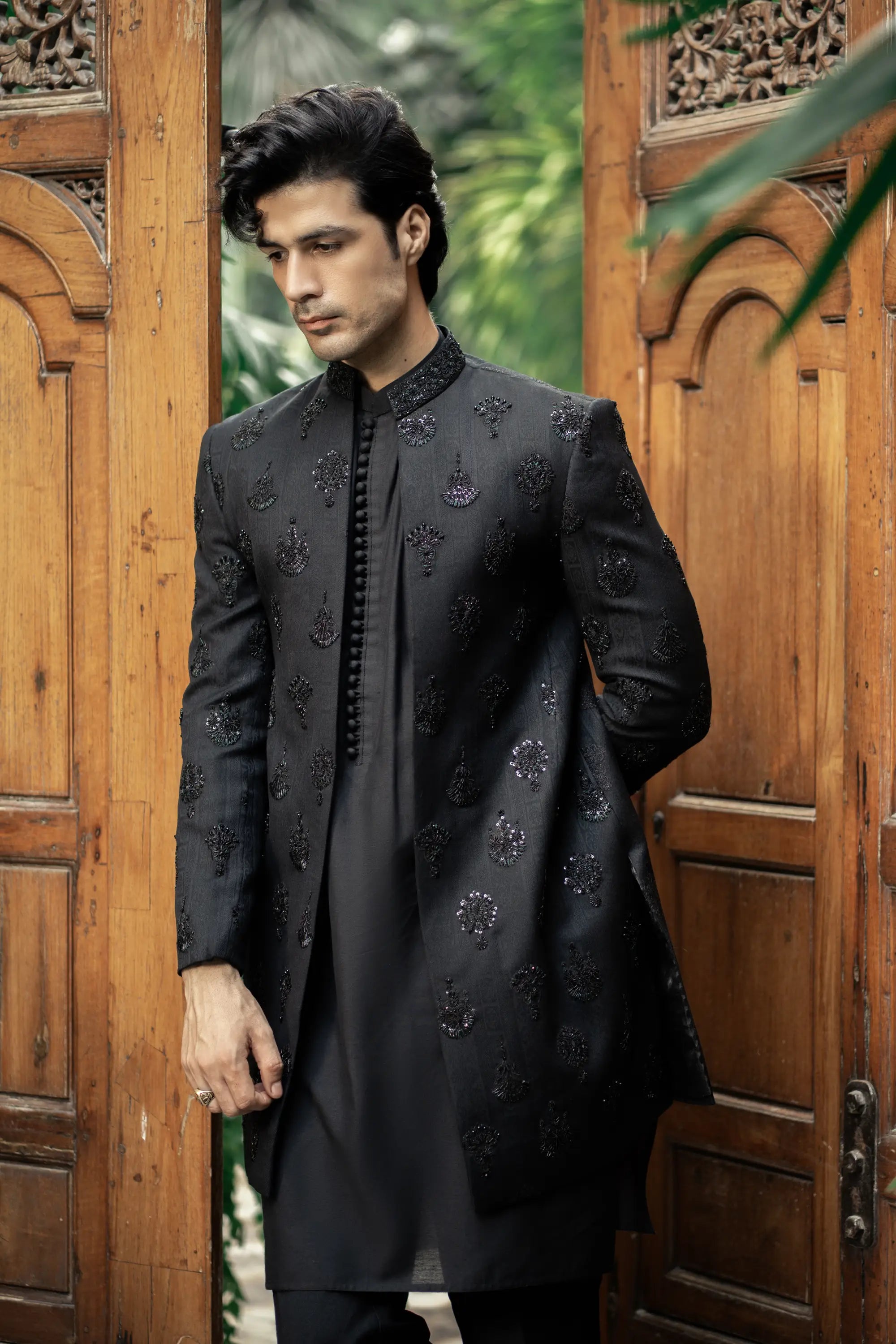 Man wearing a black embroidered sherwani standing in front of wooden doors with greenery in the background