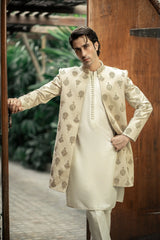 Man wearing a cream sherwani with intricate patterns standing against a wooden door.
