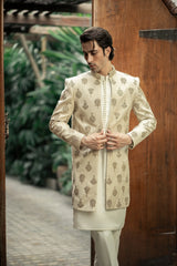 Man wearing a traditional cream sherwani with intricate patterns against a wooden door and green foliage background.