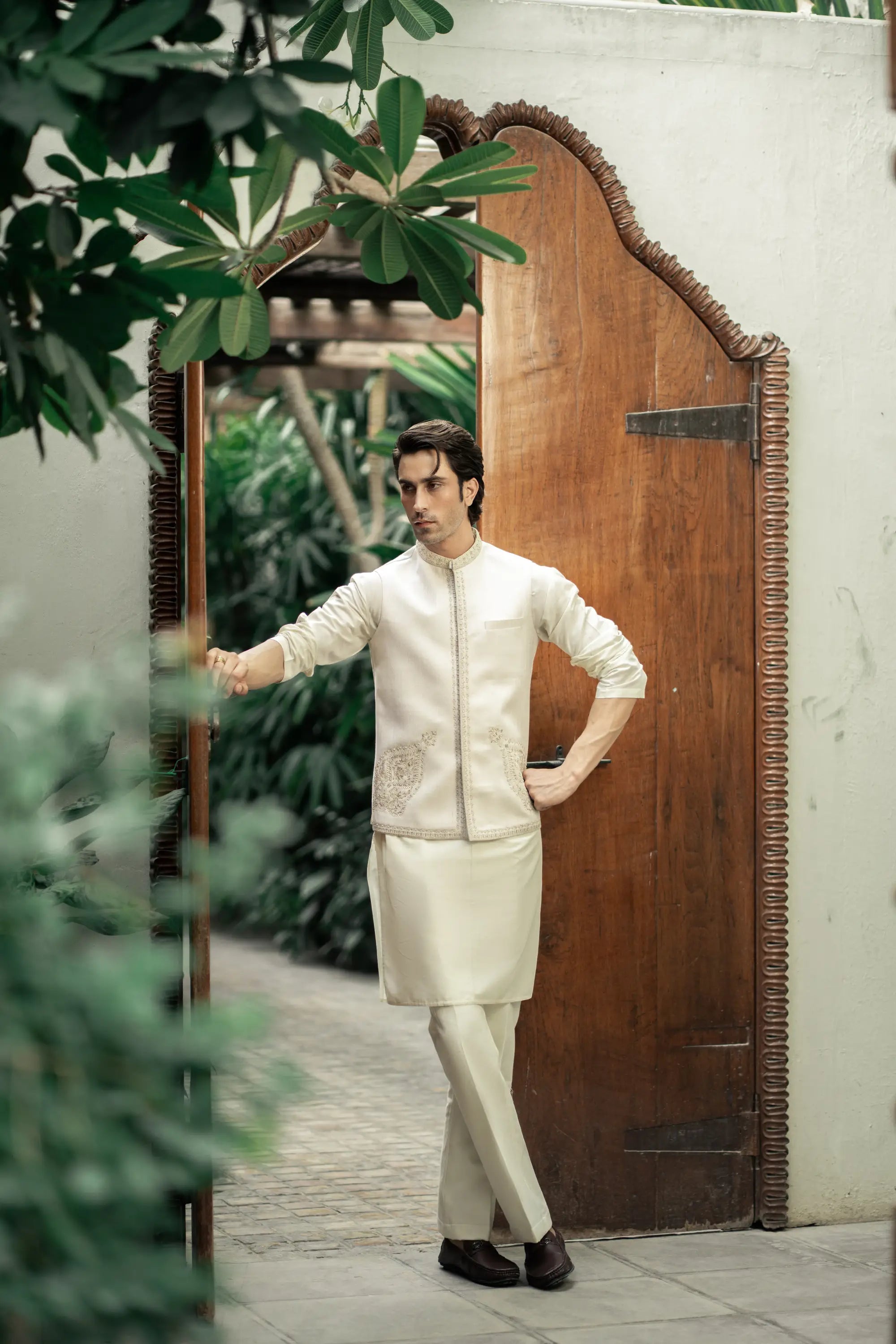 Man in a cream-colored traditional outfit standing in front of a wooden door with plants around.