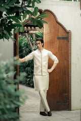 Man in a cream-colored traditional outfit standing in front of a wooden door with plants around.