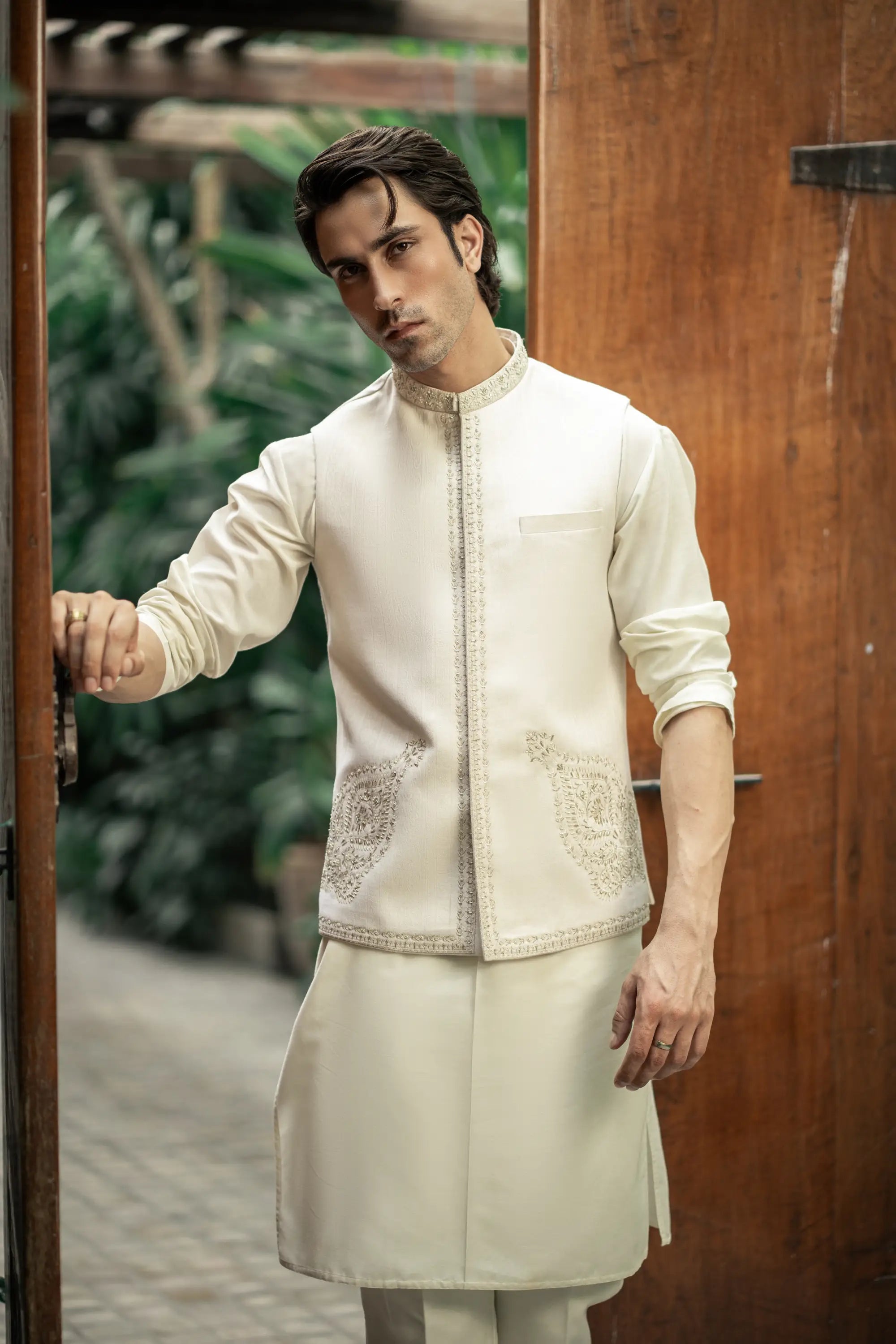 Man wearing a traditional cream kurta with intricate patterns, standing in front of a wooden door.