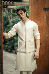 Man wearing a traditional cream kurta with intricate patterns, standing in front of a wooden door.