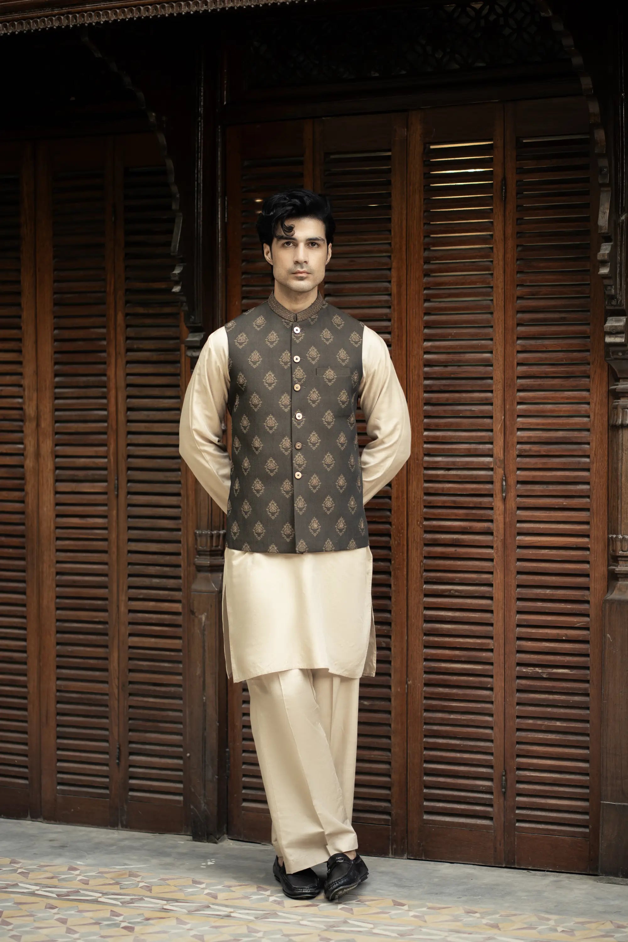 Man wearing a traditional outfit with a vest in front of wooden shutters