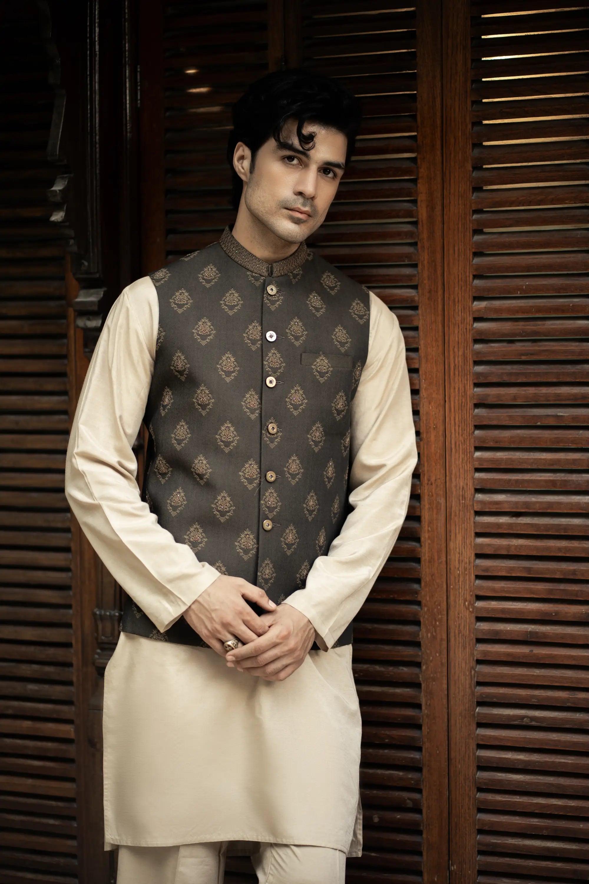 Man wearing a traditional outfit with a dark vest over a cream kurta against a wooden background
