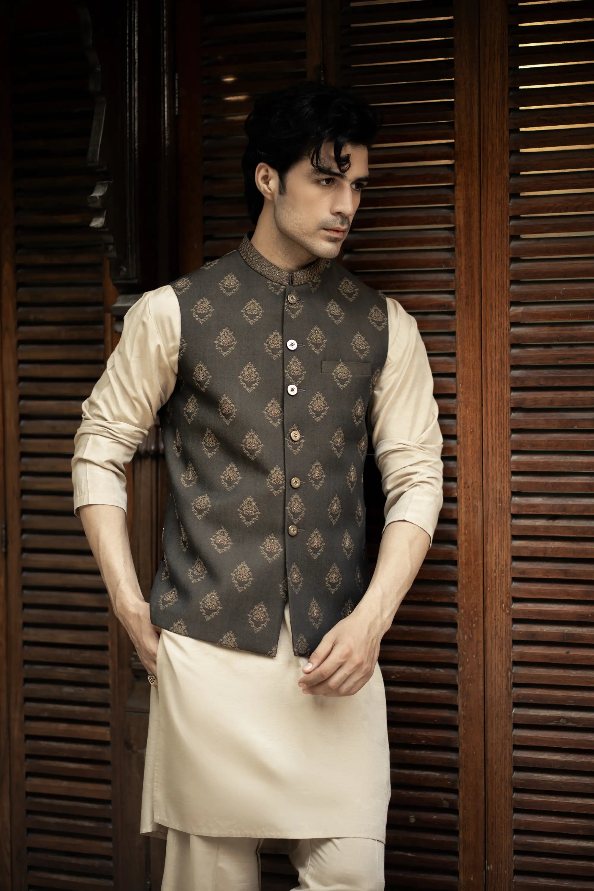 Man wearing a traditional dark gray vest over a cream kurta against a wooden background