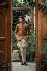 Man in a patterned jacket and light pants standing between wooden doors with greenery in the background