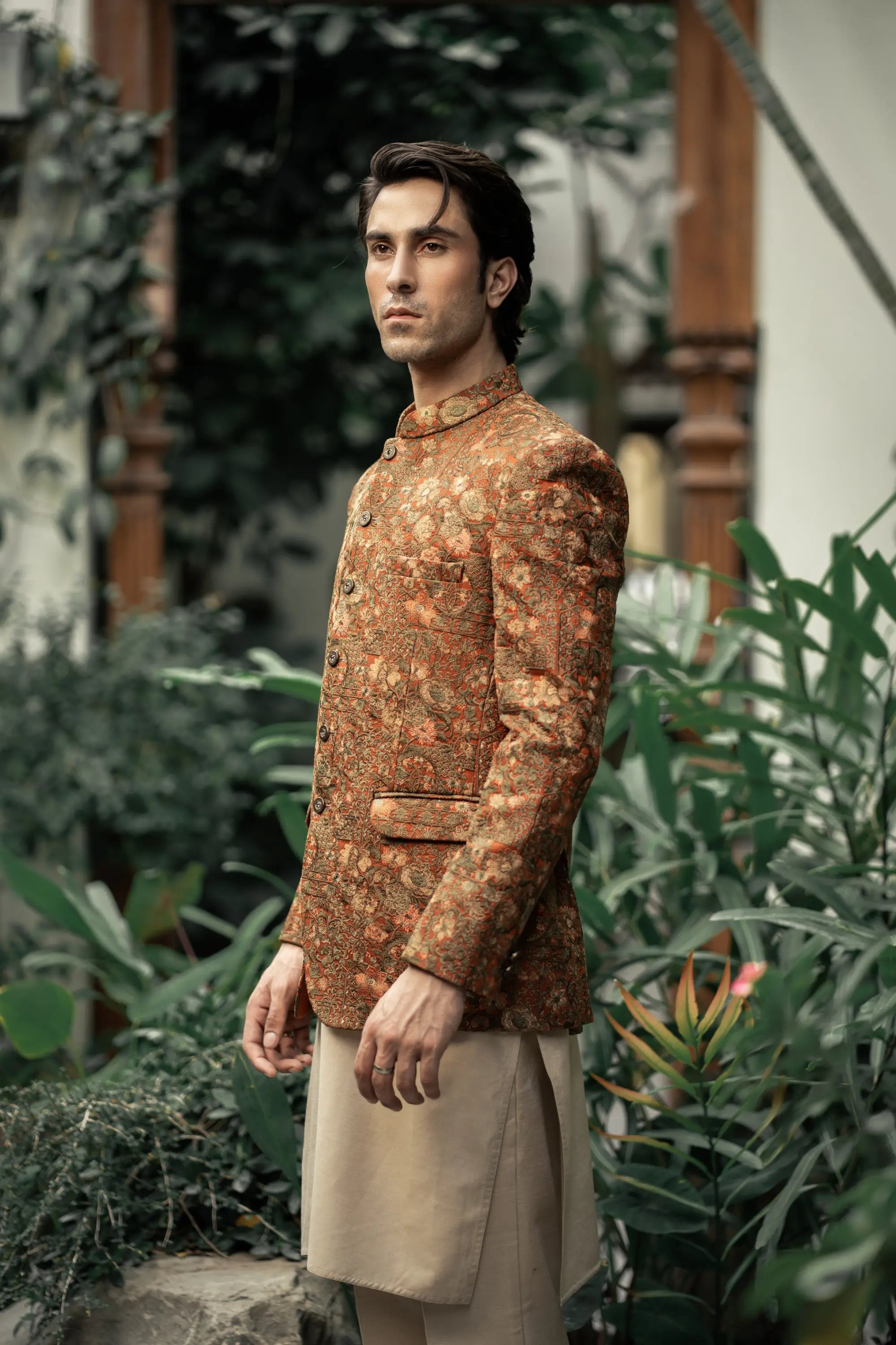 Man wearing a patterned jacket and beige pants standing in a garden setting.