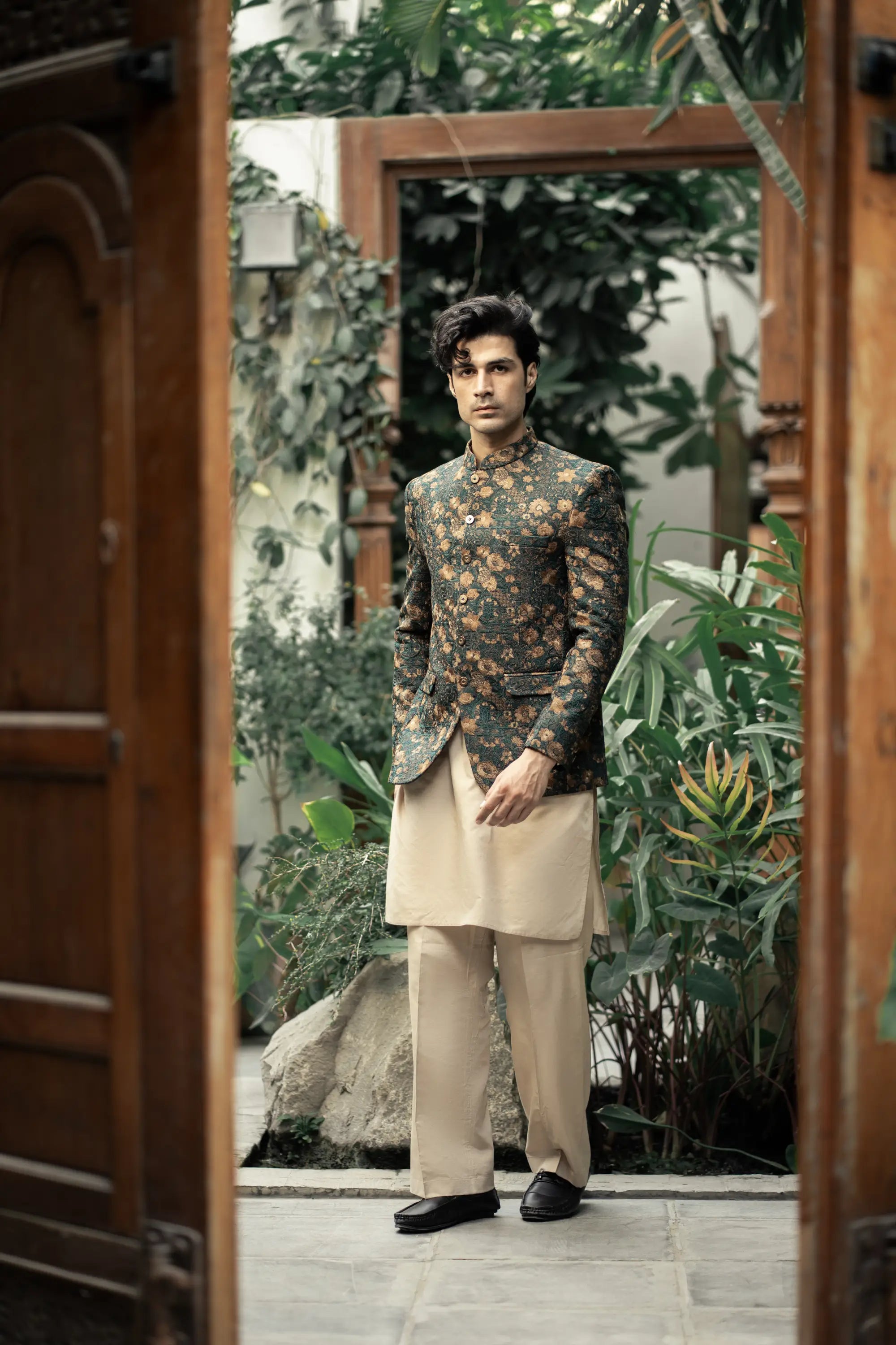Man in a floral jacket and beige pants standing in a doorway with greenery outside.
