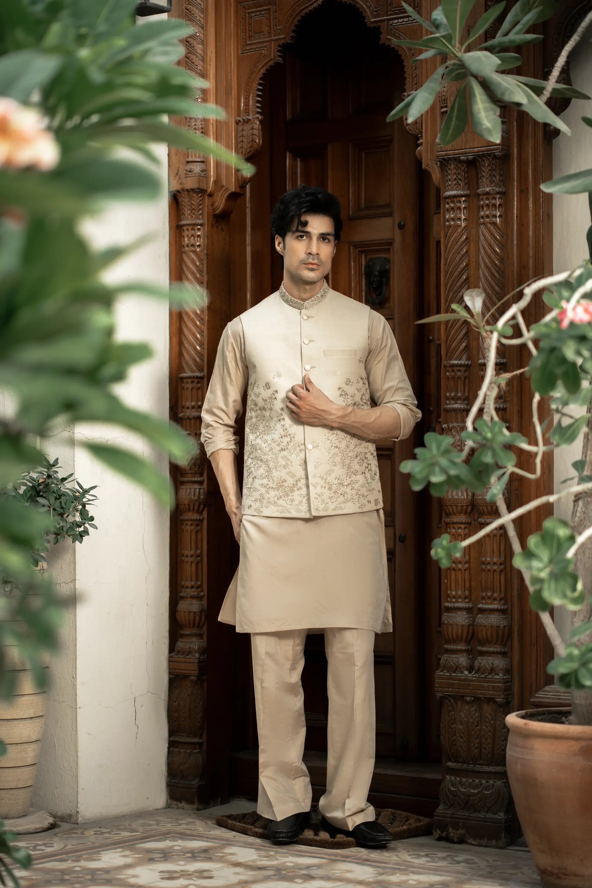 Man in traditional beige kurta standing in front of a wooden door with plants around.