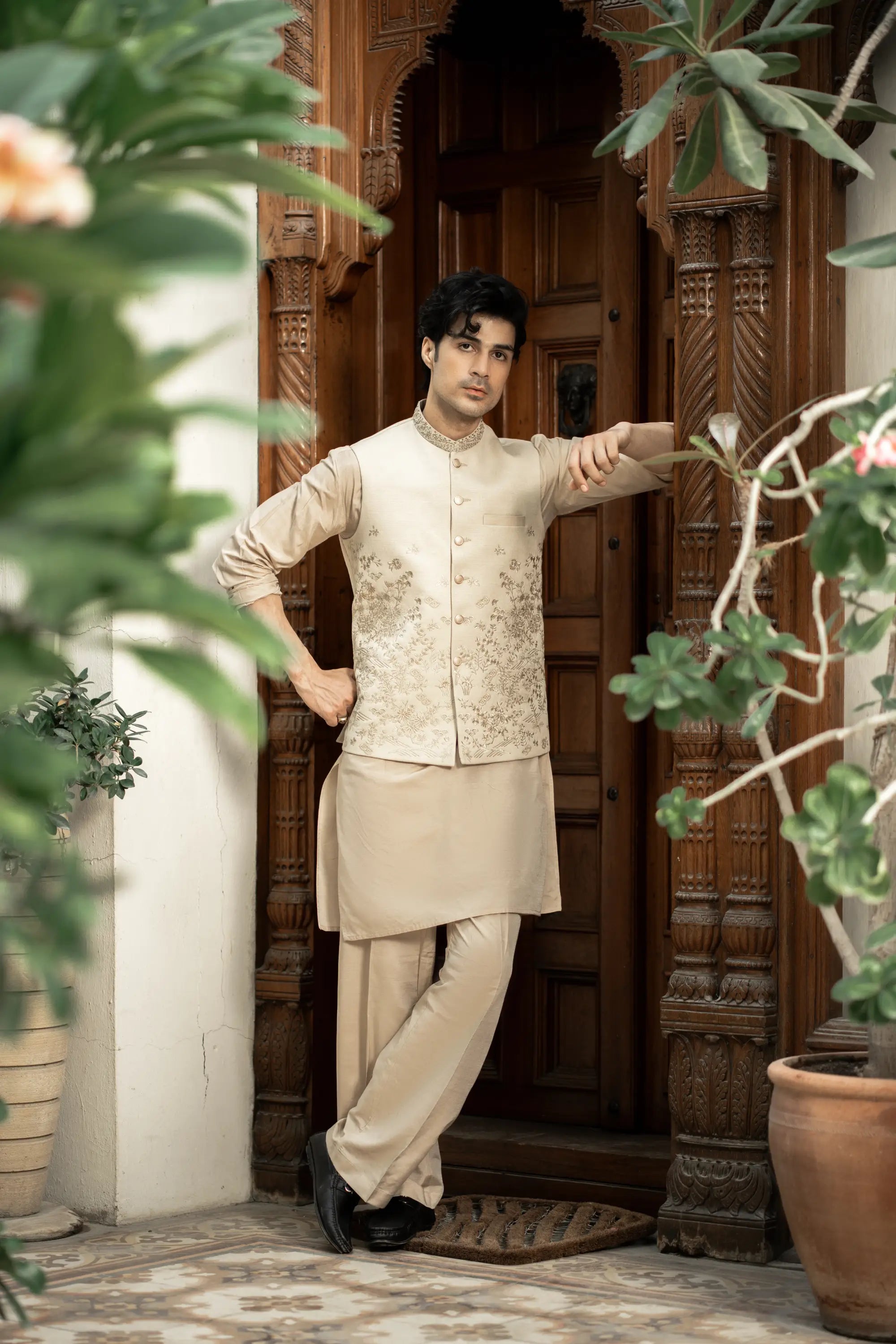 Man in traditional beige outfit standing in front of a wooden door with plants around.